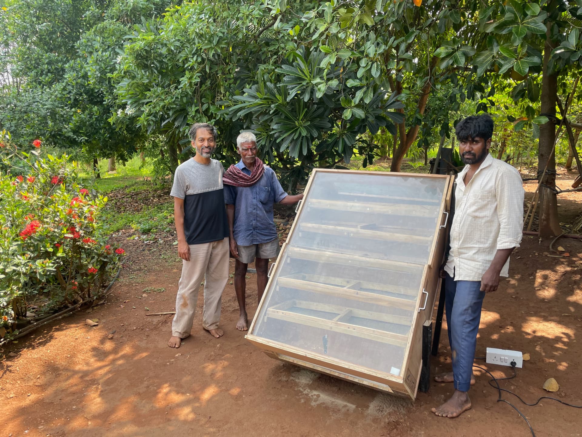 Three men standing beside a wooden solar dryer under the trees